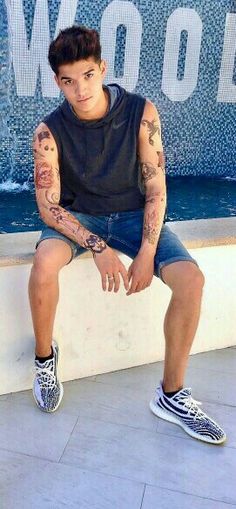 a young man with tattoos sitting on a ledge next to a fountain and posing for the camera