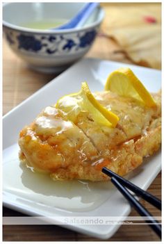 a white plate topped with chicken covered in gravy and lemon wedges next to chopsticks