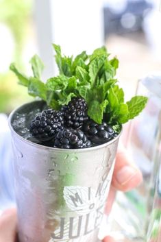 a person holding a metal cup filled with blackberries and green leaves on top of it