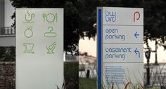 two white signs with green lettering on them in front of some trees and grass near the water