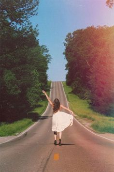 a woman is walking down the road with her arms in the air and trees behind her