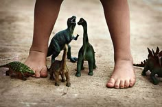 three toy dinosaurs standing next to a child's feet with one dinosaur on the ground