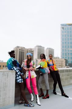 four women in colorful outfits standing on a bridge with tall buildings behind them and one woman leaning against the wall