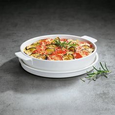 a casserole dish with vegetables in it on a gray tablecloth next to a sprig of rosemary