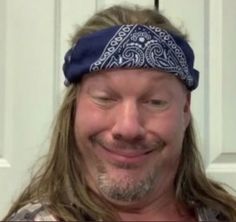 a man with long hair wearing a bandana smiles at the camera while standing in front of a white door