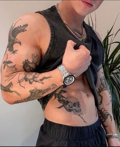 a man with tattoos on his arm and chest holding a watch in one hand while standing next to a potted plant