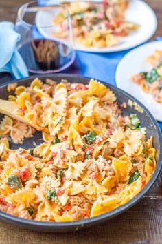 a skillet filled with pasta and vegetables on top of a wooden table next to plates