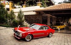 an old red car parked in front of a house next to another yellow car on the driveway