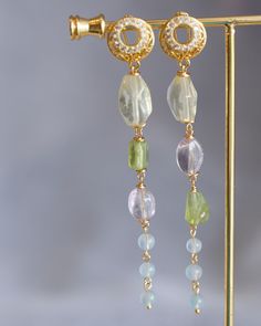 Crafted from the finest materials, these elegant earrings are a charming combination of 14K gold filled wires and a selection of luxurious gemstones, including Lemon Quartz, Amethyst, Peridot, and Aquamarine. Suspended from a pair of cubic ear hoops, they offer a unique accent to your everyday wardrobe or a thoughtful gift for someone special. Images cannot capture their beauty - you must see them in person to appreciate their grandeur. Follow me on Instagram: @cubic_jewels Elegant 14k Gold-filled Jewelry With Gemstone Accents, Dainty Gold Earrings With Gemstone Accents, 14k Gold-filled Yellow Gold Earrings With Natural Stones, Gold Dangle Gemstone Linear Earrings, Gold Dangle Crystal Earrings With Gemstone Accents, Gold Plated Earrings With Gemstone Accents, Gold-plated Earrings With Gemstone Accents, Gold Long Drop Jewelry With Gemstone Accents, Elegant Wire Wrapped Briolette Earrings