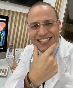 a man wearing glasses smiles while sitting in front of a computer monitor and cell phone