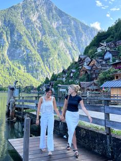 two women are walking on a dock in front of a mountain village with houses and mountains behind them