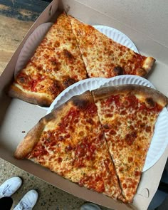 two slices of pizza sitting in a box with paper plates on the floor next to it