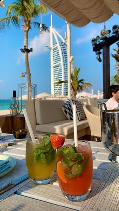 two drinks sitting on top of a wooden table next to the ocean and palm trees