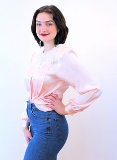 "This is a sweet vintage 80s lace collar blouse! It's made of shimmery blush pink-peach satin, very lustrous, with modest pleating in the front, puffed shoulders with shoulder pads, and long sleeves with single button cuffs. It has a very pretty, large white cut lace collar, a round neckline, and it closes with six white pearl buttons in a hidden front placket, plus a snap at the throat. The blouse is in very good clean vintage condition, no flaws noted. The label says \"Nicola, 100% Polyester, Pink Satin Feminine Blouse, Pink Satin Blouse In Feminine Style, Feminine Fitted Peach Blouse, Fitted Feminine Peach Blouse, Feminine Pink Satin Top, Chic Pink Blouse With Lace Collar, Vintage Satin Tops For Spring, Feminine Pink Blouse With Lace Top, Fitted Pink Blouse With Lace Collar