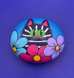 a painted rock with flowers and a cat's face on it, against a purple background