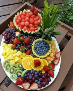 a plate full of fruit and vegetables on a wooden table with plants in the background