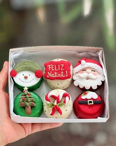 a hand holding an open box of christmas cupcakes with frosting and decorations