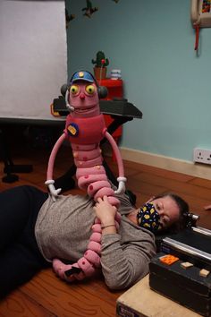 a man laying on the floor next to a giant pink object with eyes and legs