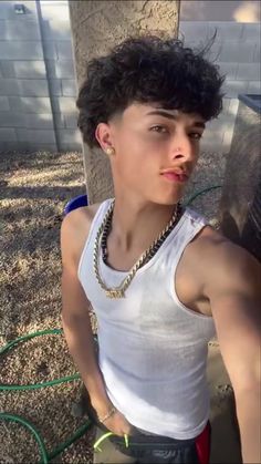 a young man wearing a white tank top standing next to a tree with chains on his neck