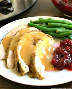 a white plate topped with meat covered in gravy next to green beans and cranberry sauce