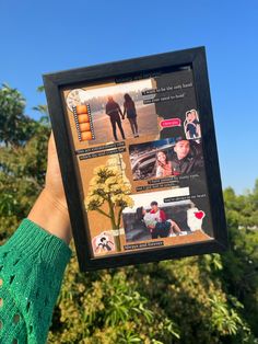 a person holding up a framed photo in front of some trees and bushes with pictures on it