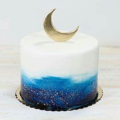 a white and blue cake with a gold crescent decoration on top, sitting on a wooden table