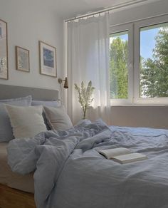 an unmade bed with blue sheets and pillows in front of two windows that have trees outside