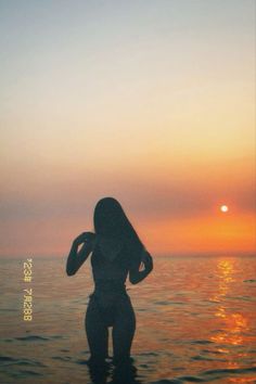 a woman standing in the water at sunset with her back to the camera and arms behind her head