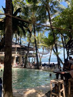 people sitting at tables next to a pool in the middle of palm trees and water