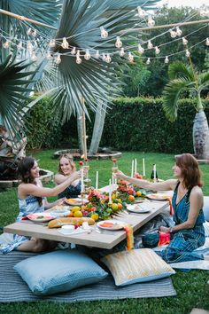 three women sitting at a table with food and drinks in front of them, surrounded by palm trees