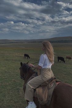 a woman riding on the back of a brown horse in a field full of cattle