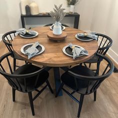 a round wooden table with black chairs and plates on it in front of a white vase