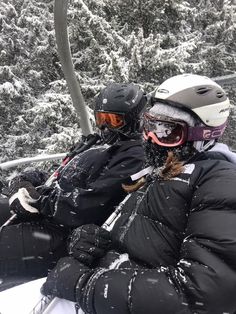 two people sitting on a ski lift in the snow with helmets and goggles on