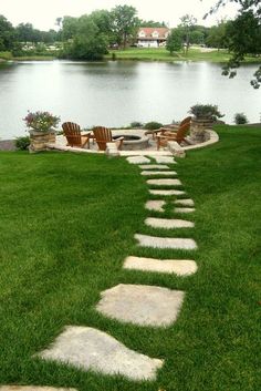 a stone path in the grass leading to a fire pit with chairs on it and a lake in the background