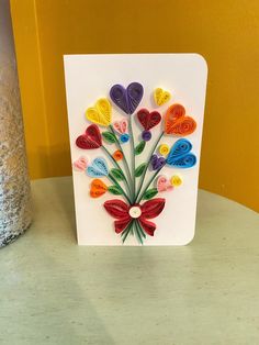 a white card with colorful paper hearts on it and a vase in the back ground