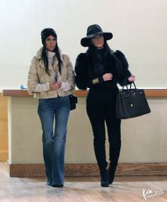 two women in hats and jackets walking down a hall way with one carrying a handbag