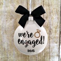 we're engaged ornament hanging on a wooden wall with a black ribbon