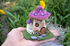 a hand holding a tiny mushroom house in the middle of some grass and flowers behind it