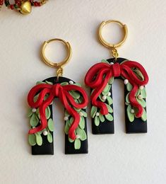 three pairs of earrings with red bows and green leaves hanging from gold hoops on a white surface