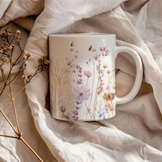 a coffee cup with flowers painted on it sitting on a white blanket next to dried plants