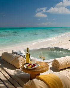 a hot tub sitting on top of a sandy beach next to the ocean with two glasses of wine