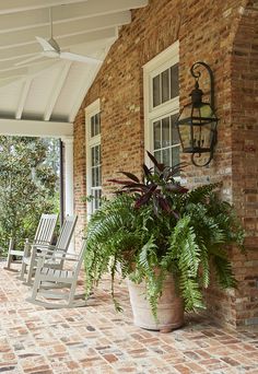 a porch with rocking chairs and a potted plant on the front steps, next to a brick wall