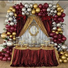 a table topped with lots of silver and gold balloons next to a red drapes