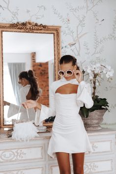 a woman standing in front of a mirror wearing a white dress and holding up her sunglasses