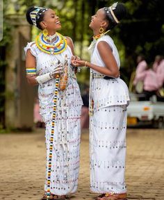 two women dressed in white standing next to each other on a brick ground with trees in the background