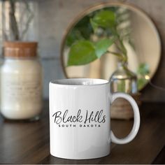 a white coffee mug sitting on top of a wooden table next to a potted plant