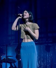 a woman standing on top of a stage with a microphone in her hand and wearing a blue skirt