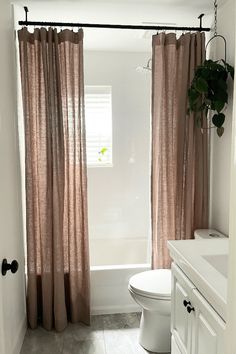 a white toilet sitting next to a bath tub in a bathroom under a window with pink curtains