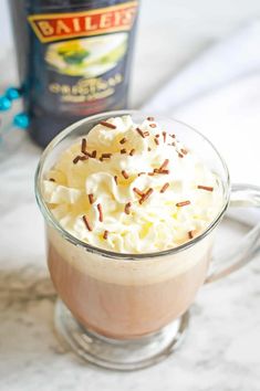 hot chocolate with whipped cream and sprinkles in a glass mug next to a bottle of bailey's