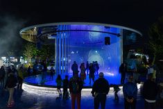 a group of people standing in front of a fountain with lights on it at night